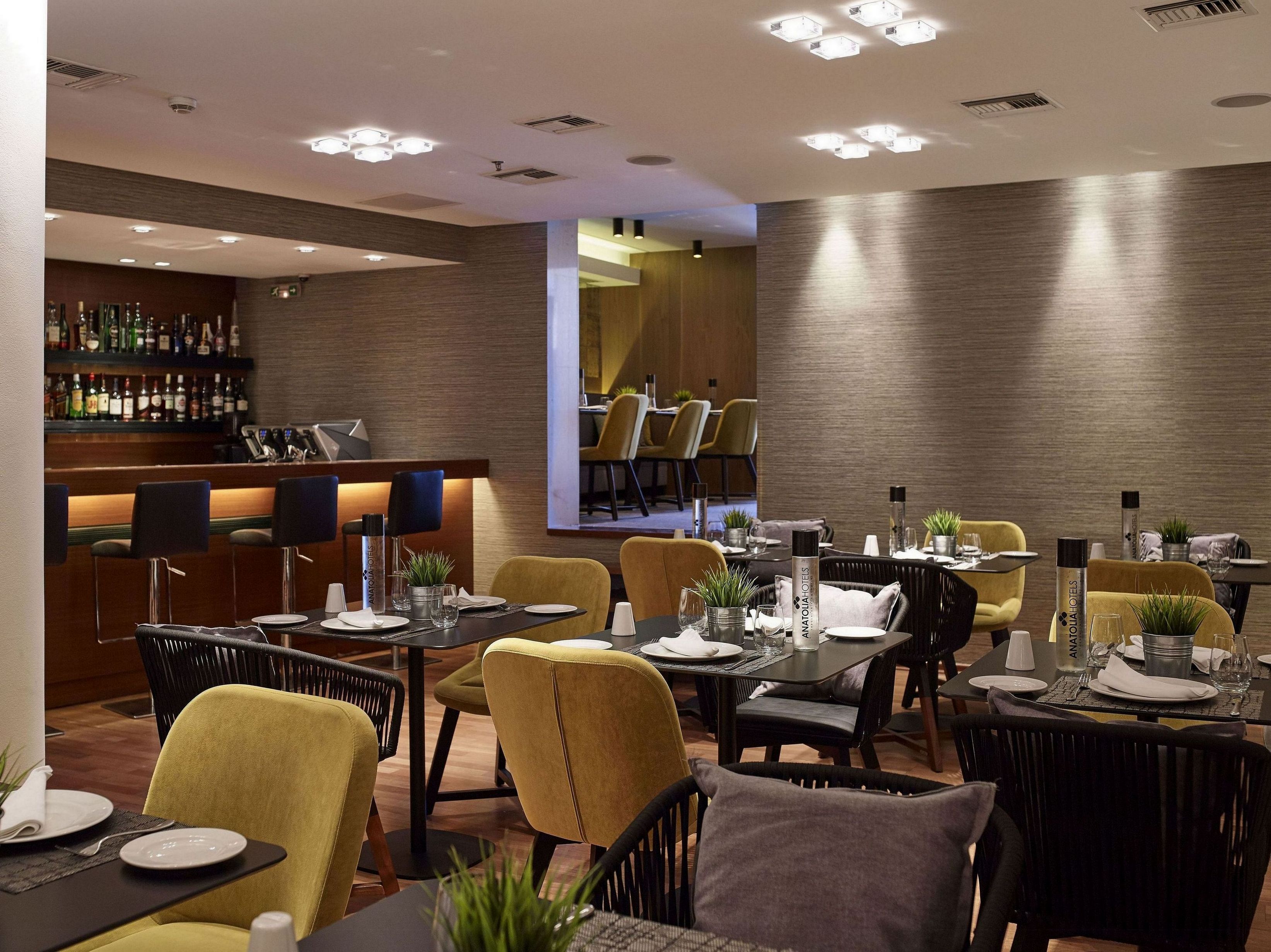 Un espace repas moderne avec des tables préparées pour deux personnes, un bar avec des bouteilles d’alcool exposées et un miroir qui reflète la pièce.
