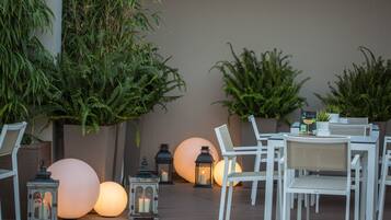 An outdoor seating area with white chairs and tables, surrounded by large potted plants and illuminated spherical lanterns.