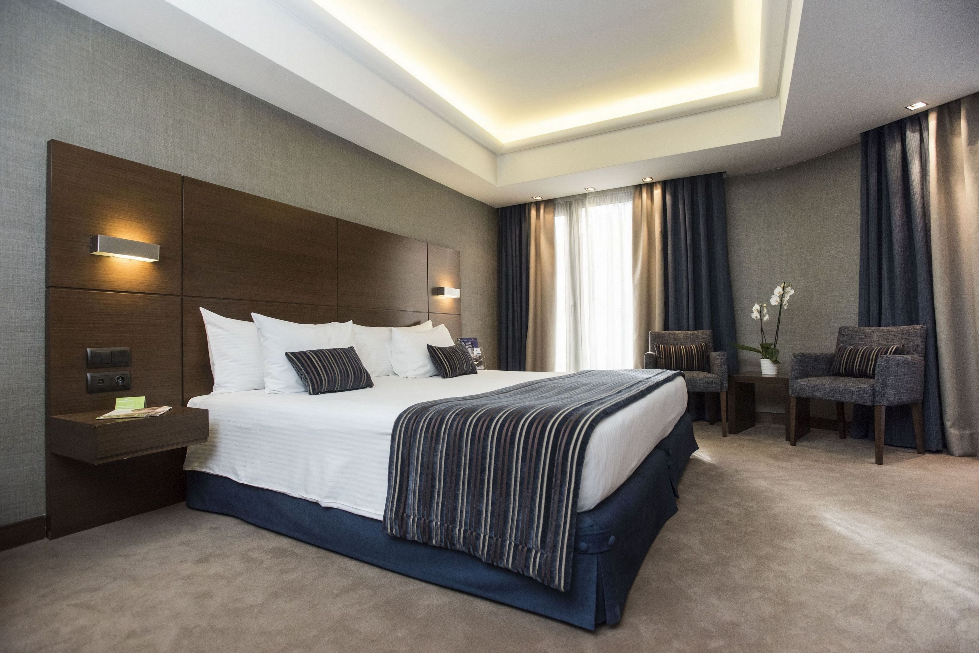 Een moderne hotelkamer met een groot bed, nachtkastjes, een zithoek met stoelen en uitzicht op het omliggende landschap.
