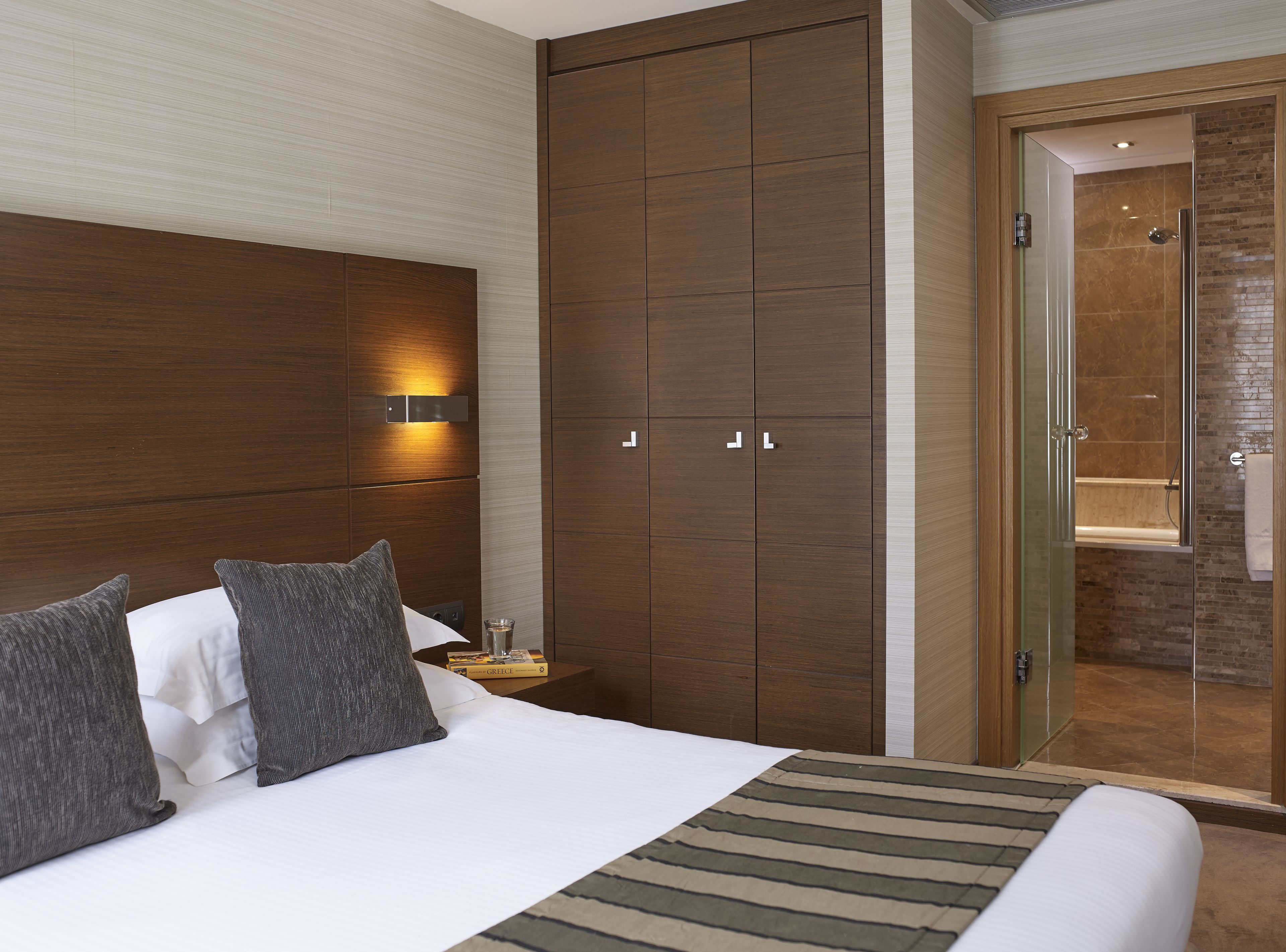 A hotel room with a large bed, a wooden wardrobe, a nightstand with a glass, and a bathroom with a glass door.