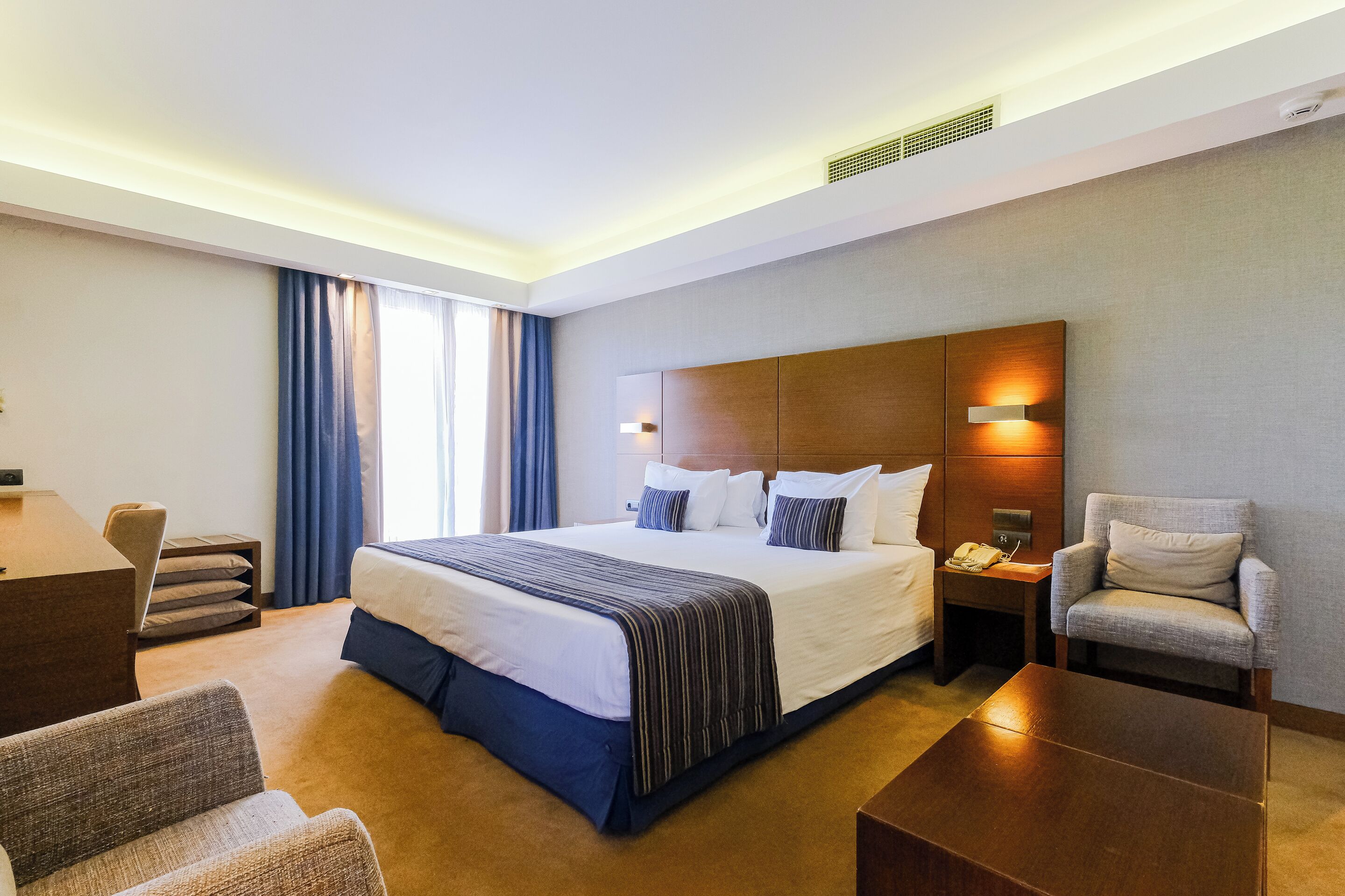 A hotel room with a large bed, bedside tables, a desk, a chair, and a sofa.