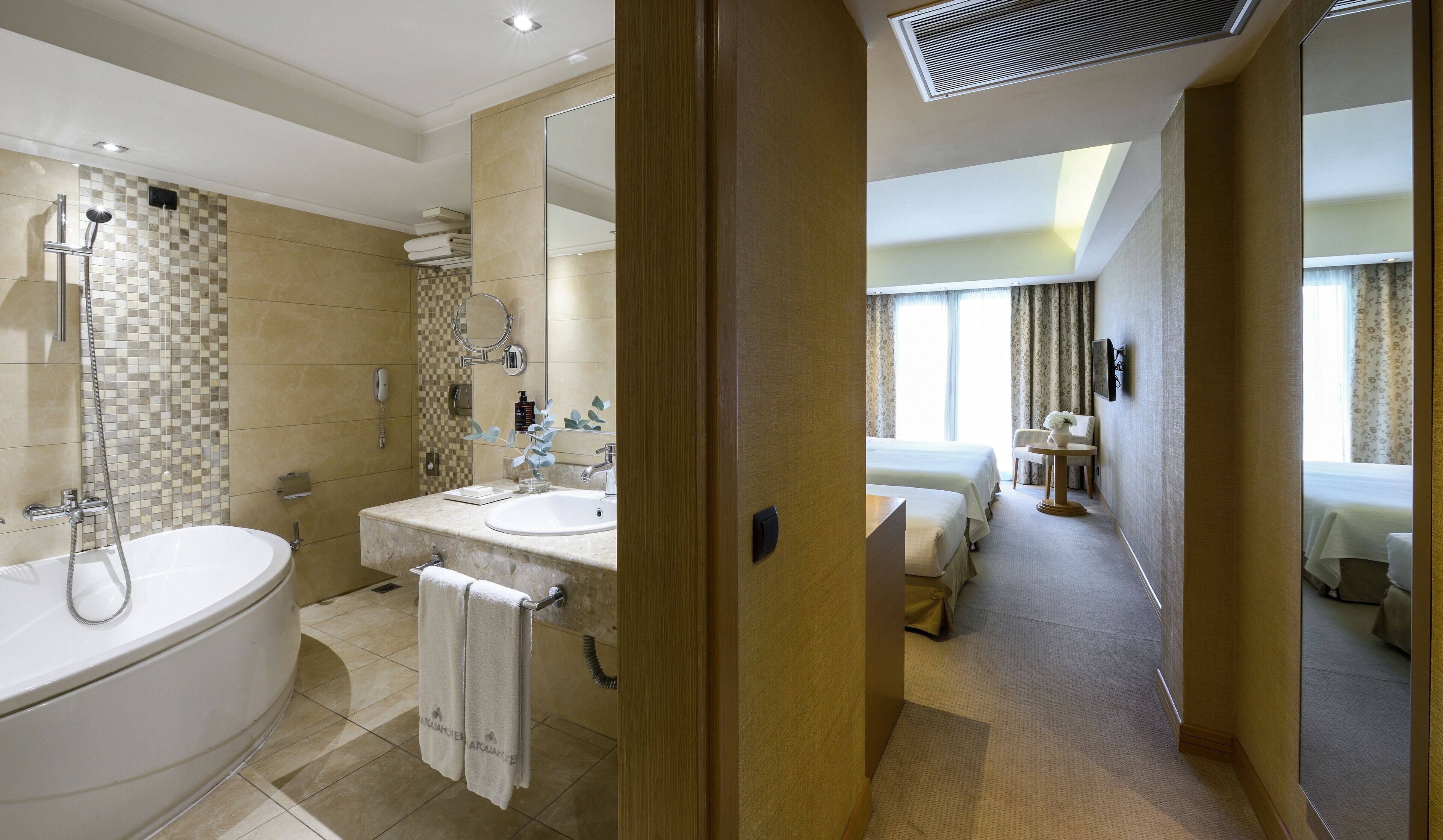 A hotel bathroom with a bathtub, two sinks, and a shower. There is a mirror above the sink and a towel rack. The bathroom has a tiled floor and a checkered wall pattern.