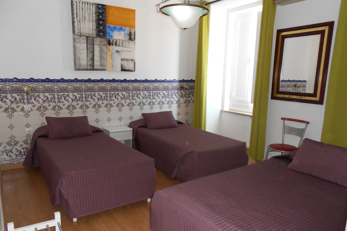 Une chambre avec deux lits simples, une petite table blanche, une chaise, une fenêtre avec des rideaux et un mur orné d’une frise de carreaux décoratifs.