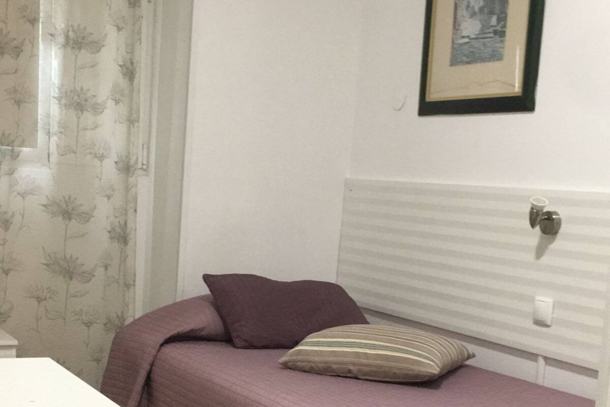 Une petite pièce comprenant un lit, une table de chevet, une fenêtre avec des rideaux et un tableau encadré accroché au mur.