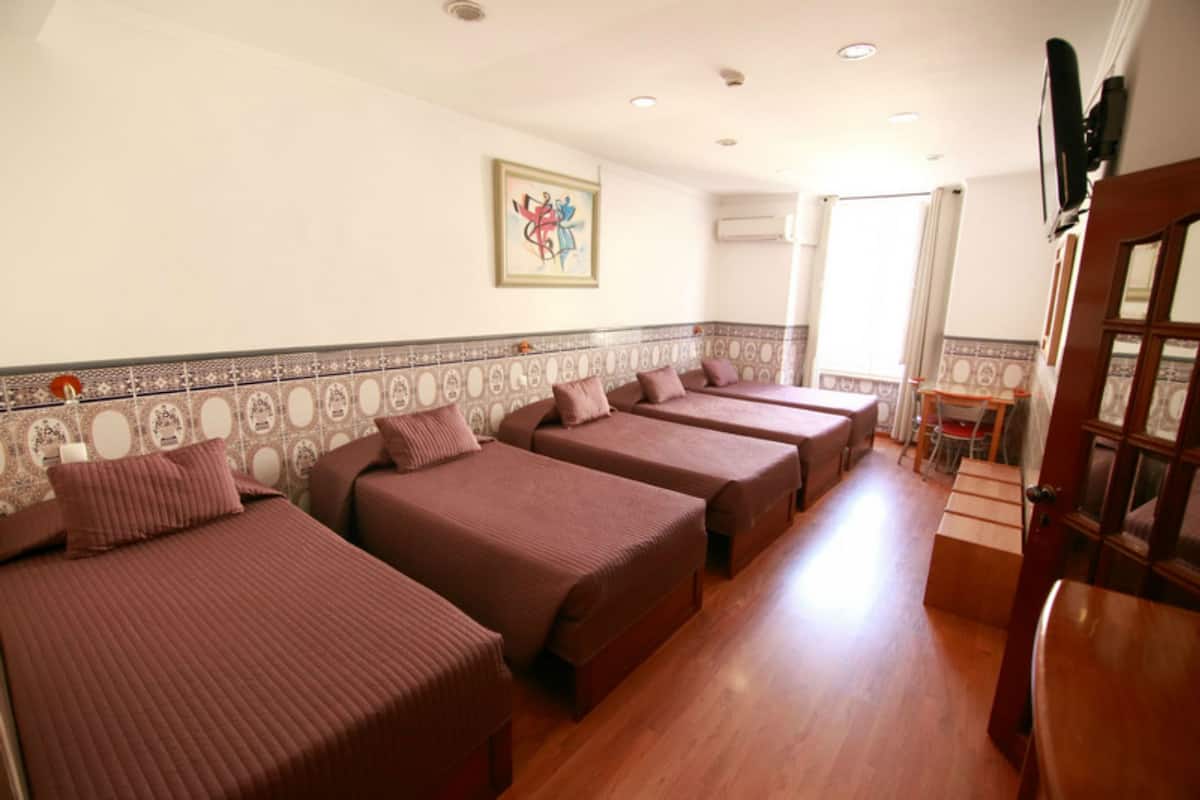Une chambre d’hôtel avec quatre lits simples, un plancher en bois, un mur recouvert de carreaux avec une bordure décorative, une œuvre d’art encadrée et un escalier menant à une autre pièce.
