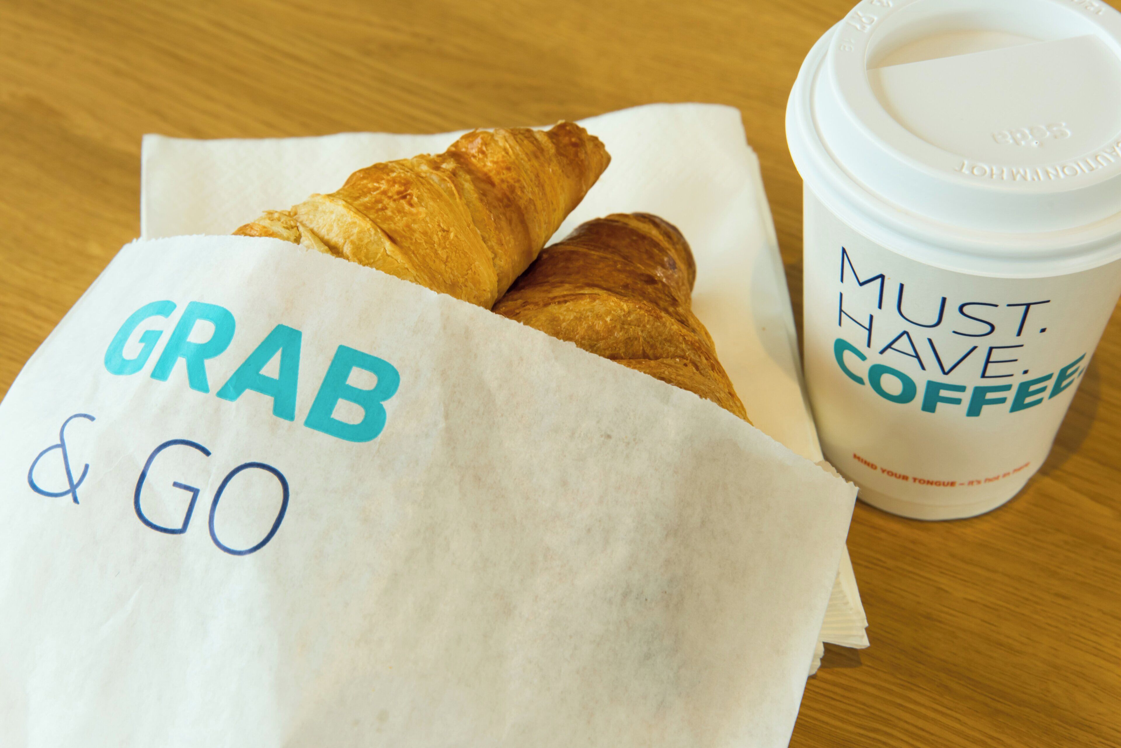 A croissant in a paper bag with the words 'GRAB & GO' and a coffee cup with the text 'MUST HAVE COFFEE'.