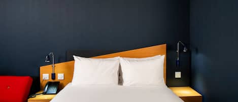 Een hotelkamer met een bed, een rode stoel, een nachtkastje met een telefoon en een wandlamp.