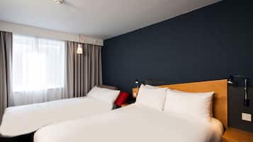 A hotel room with two beds, a window with curtains, a bedside table with a lamp, and a wall-mounted light fixture.