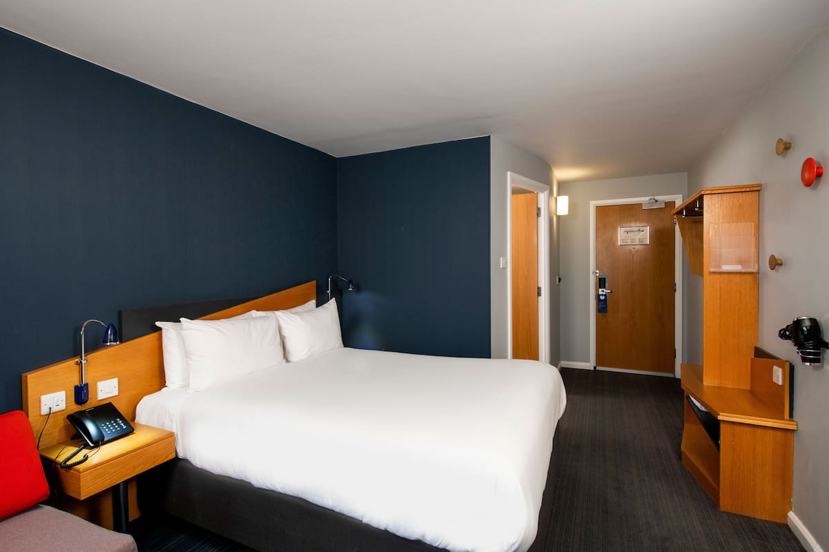 Una habitación de hotel con una cama grande, una mesita de noche con un teléfono, una silla roja y un armario de madera.