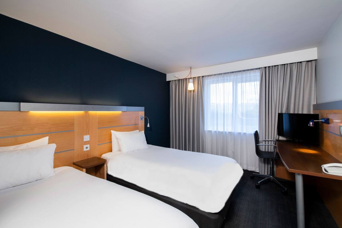 A hotel room with two beds, a desk, a television, and a window with curtains.