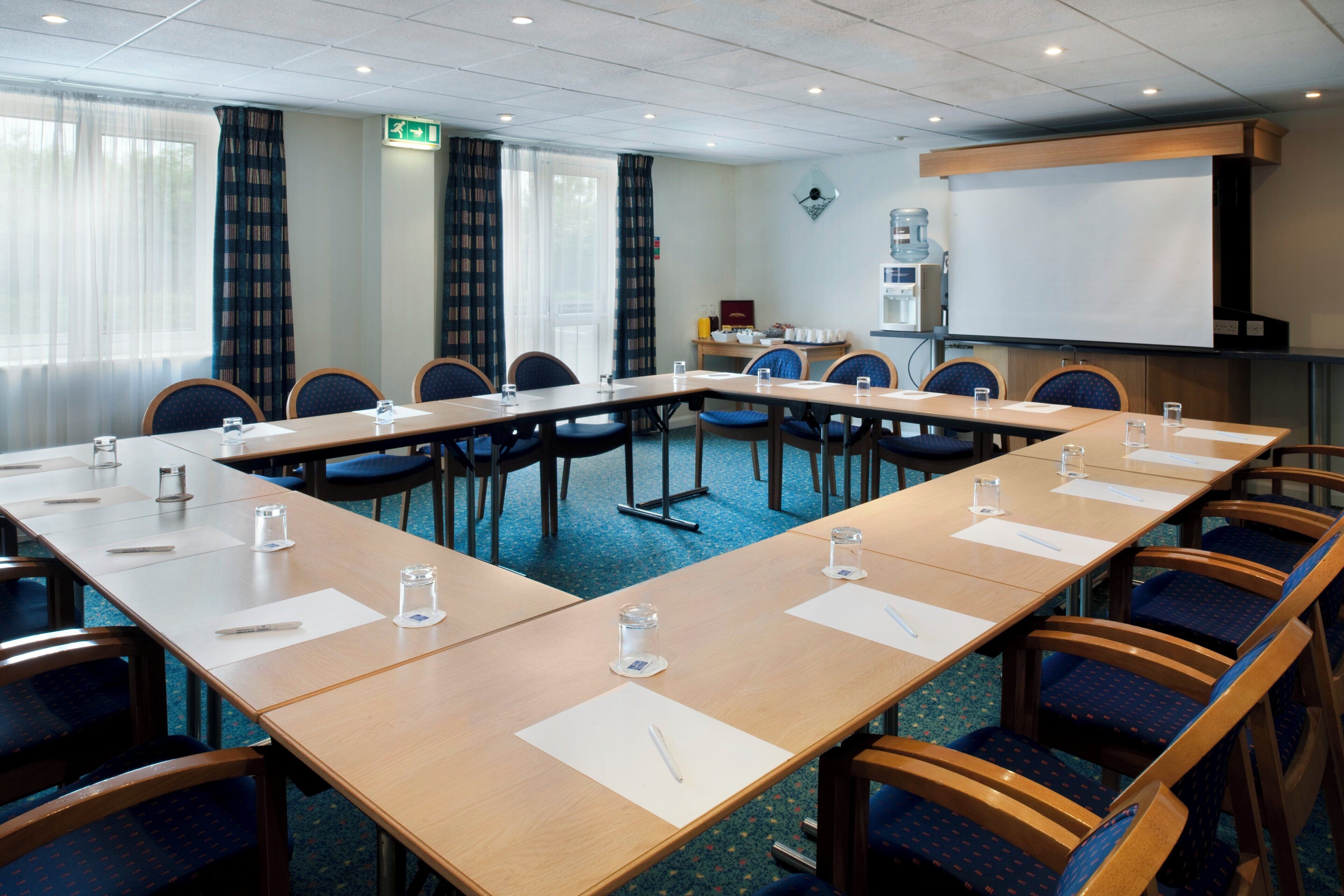 A conference room with a long table, chairs, a projector screen, and a water dispenser.