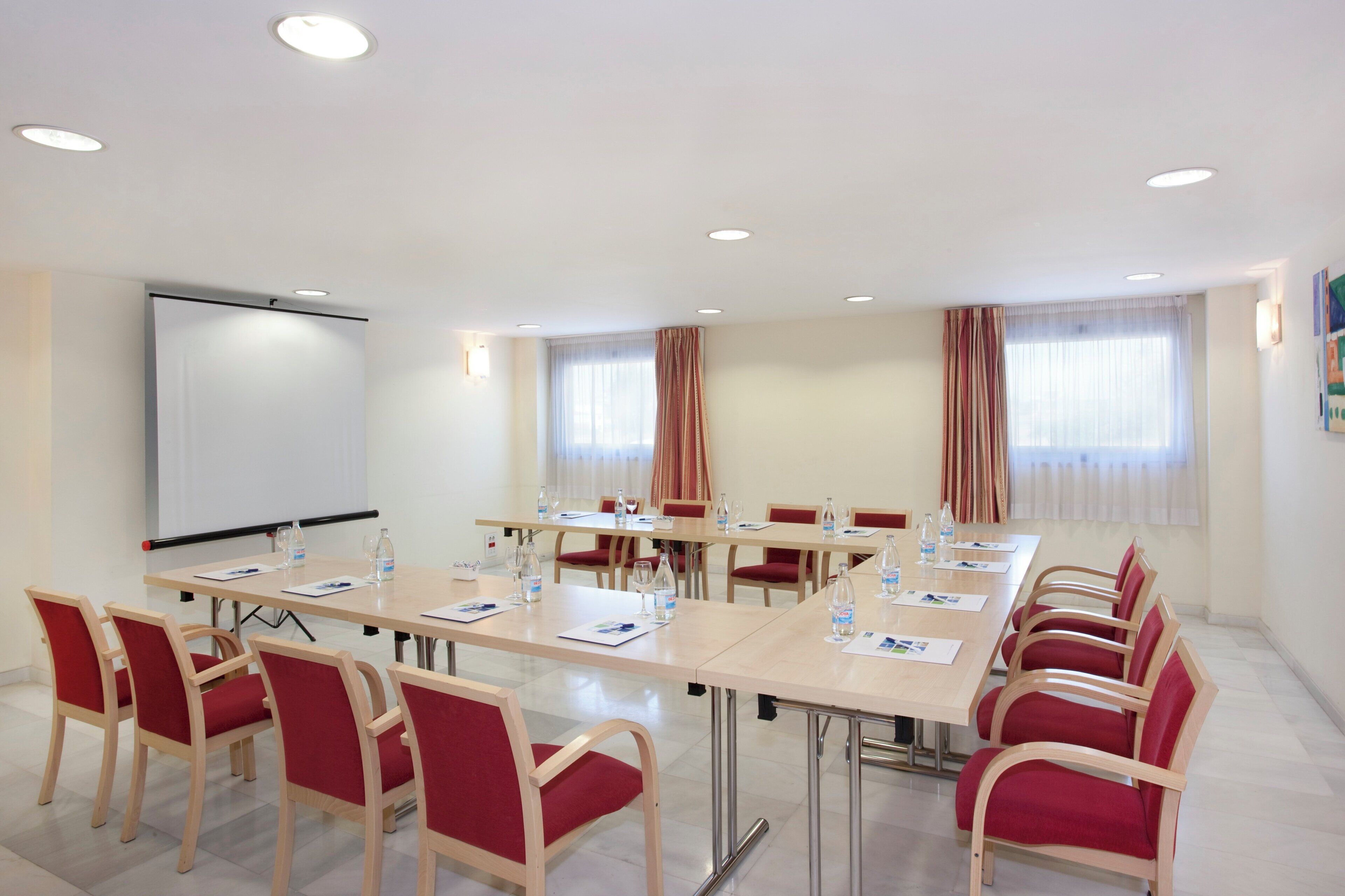 Un salón de conferencias con una gran mesa, sillas rojas, una pizarra y ventanas con cortinas.