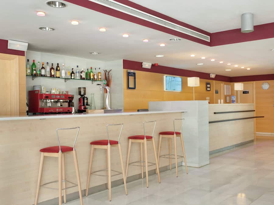 Een hotel lobby met een bar, barstoelen, een koffieapparaat en een bord met prijzen voor de kamers.