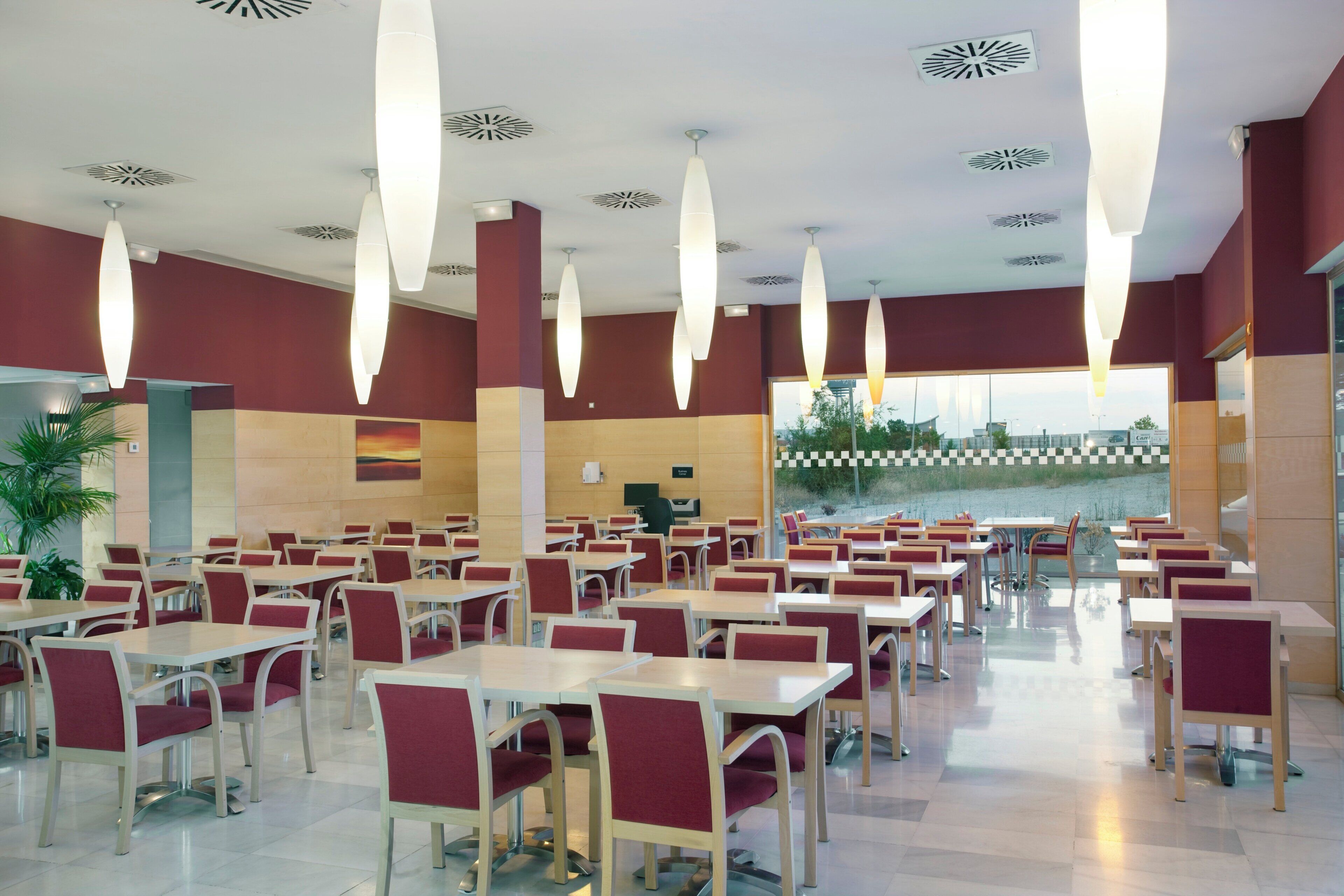 A spacious dining area with multiple tables and chairs, a large window offering a view of the outdoors, and modern ceiling fixtures.