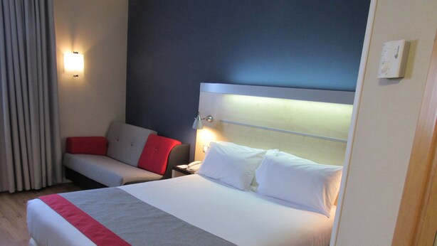 Una habitación de hotel con una cama grande, una mesita de noche con lámpara, un sofá rojo y gris, y una ventana con cortinas.