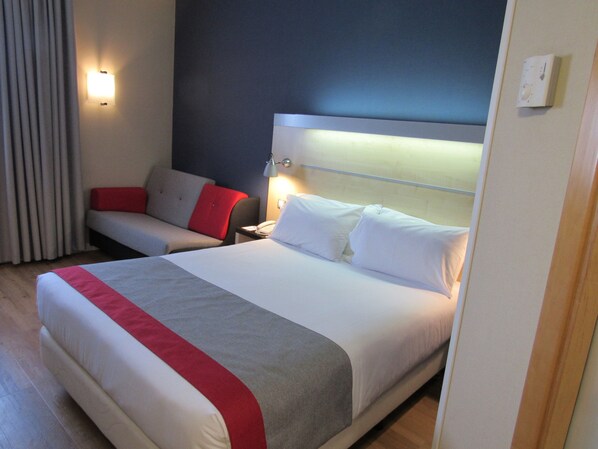 A hotel room with a large bed, a bedside table with a lamp, a red and grey sofa, and a window with curtains. - Holiday Inn Express Madrid-Alcorcón by IHG (Alcorcon)
