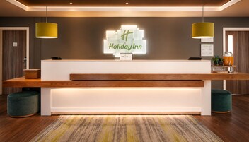 Una recepción con el letrero de Holiday Inn, mostrador de madera, dos lámparas colgantes y una planta pequeña a la derecha.