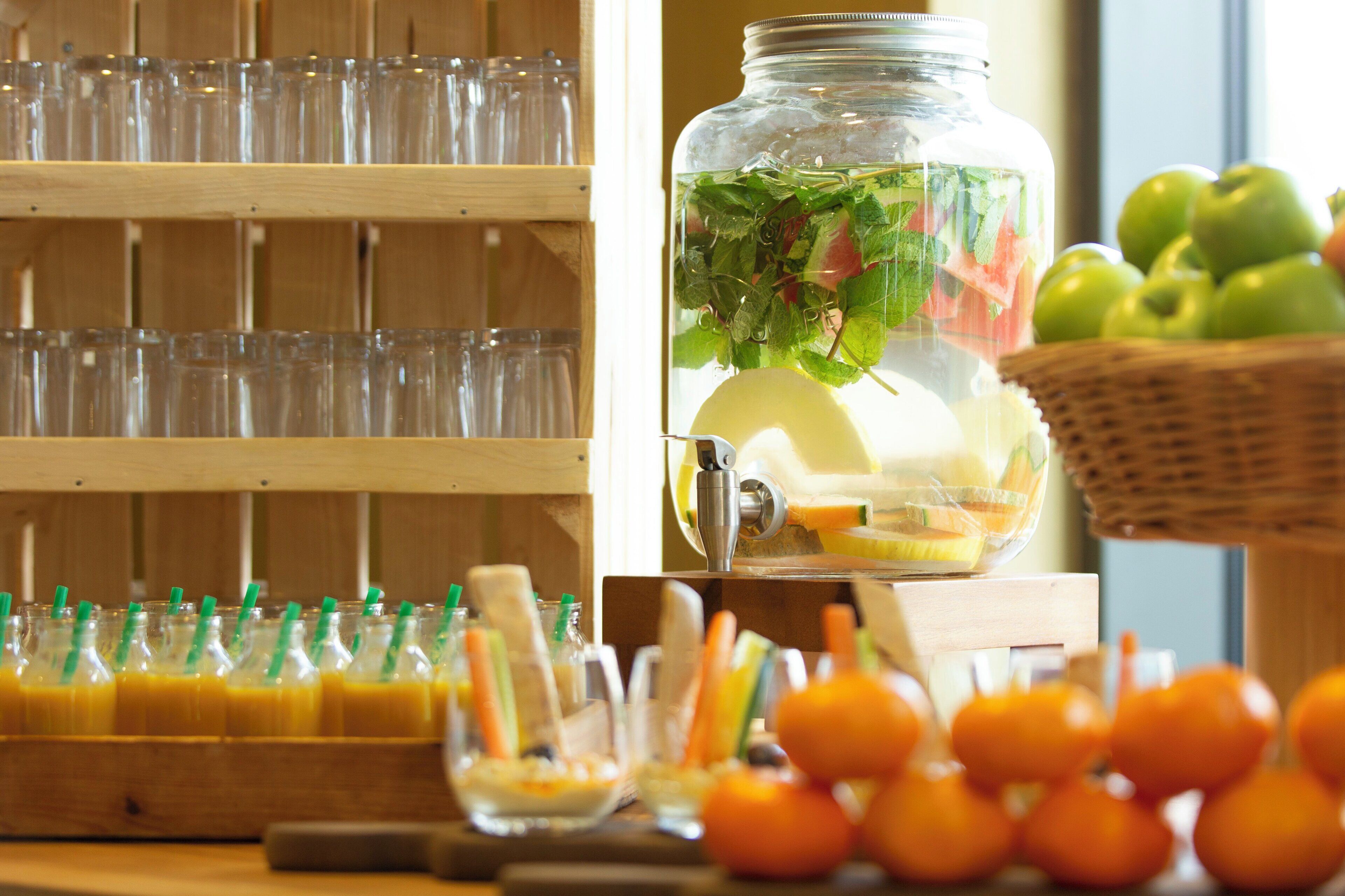 A beverage station with a water dispenser, a fruit basket, and a row of glasses.