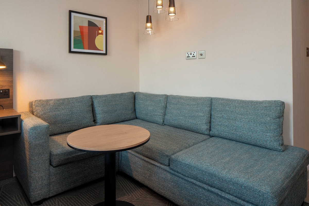 A corner sofa with a round table, a framed abstract painting, and a wall socket.