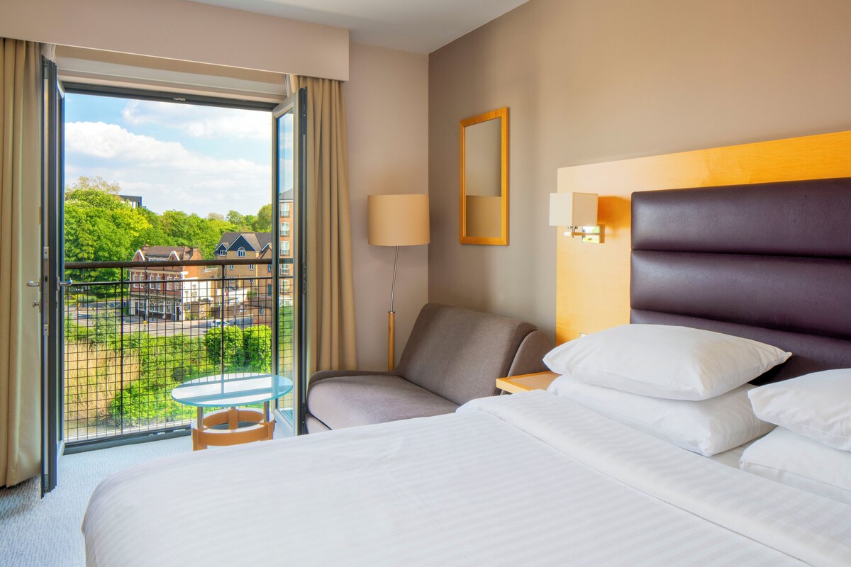 Ein Hotelzimmer mit Bett, Sessel, kleinem Tisch, Lampe, Spiegel und einem Balkon mit Blick auf Gebäude und Grünflächen.