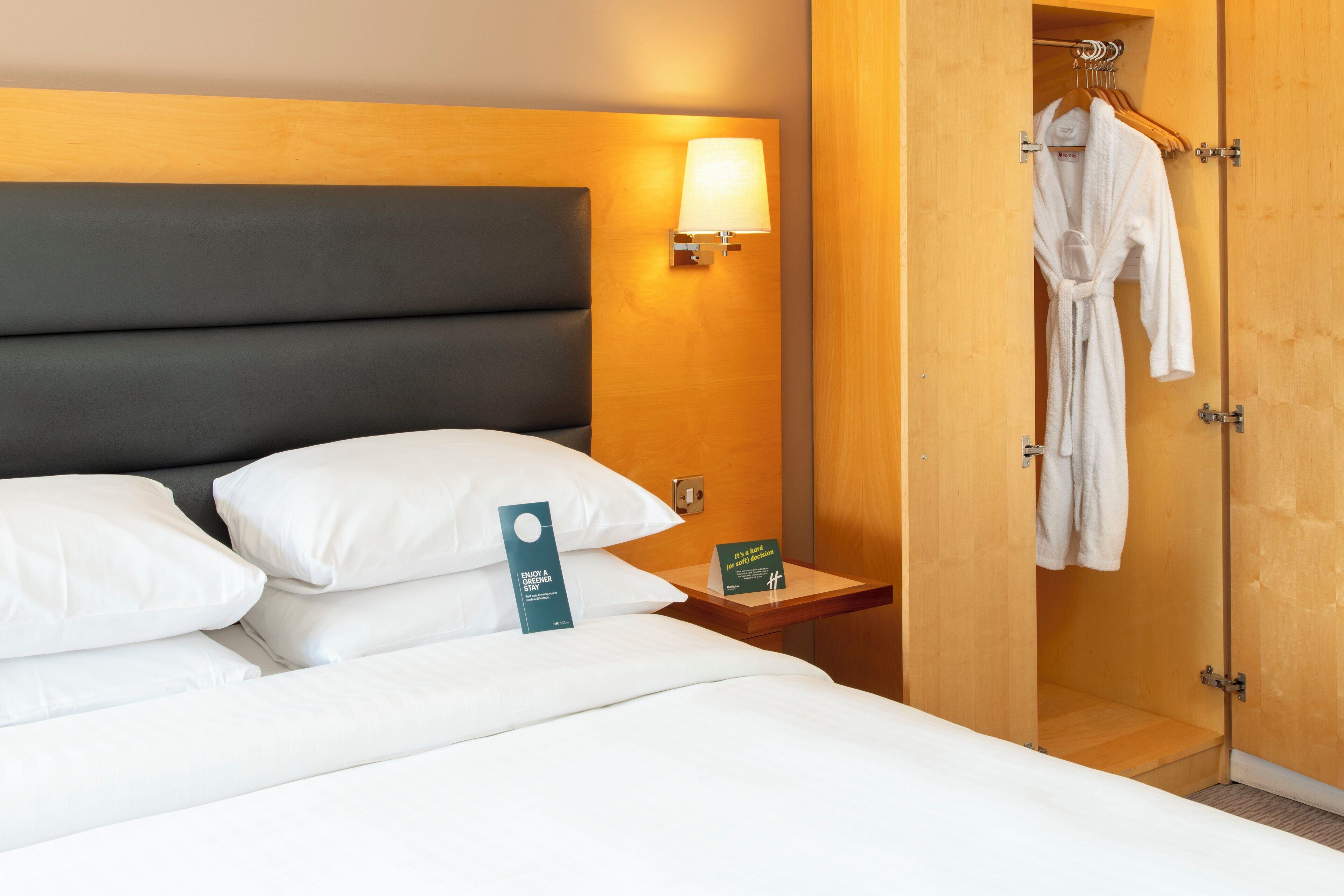 A hotel room with a bed, white pillows, a nightstand with a lamp, a robe hanging on a door, and a sign indicating the room type.