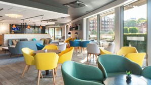 A modern lounge area with vibrant yellow and teal chairs, a wooden floor, and large windows offering a view of the city.