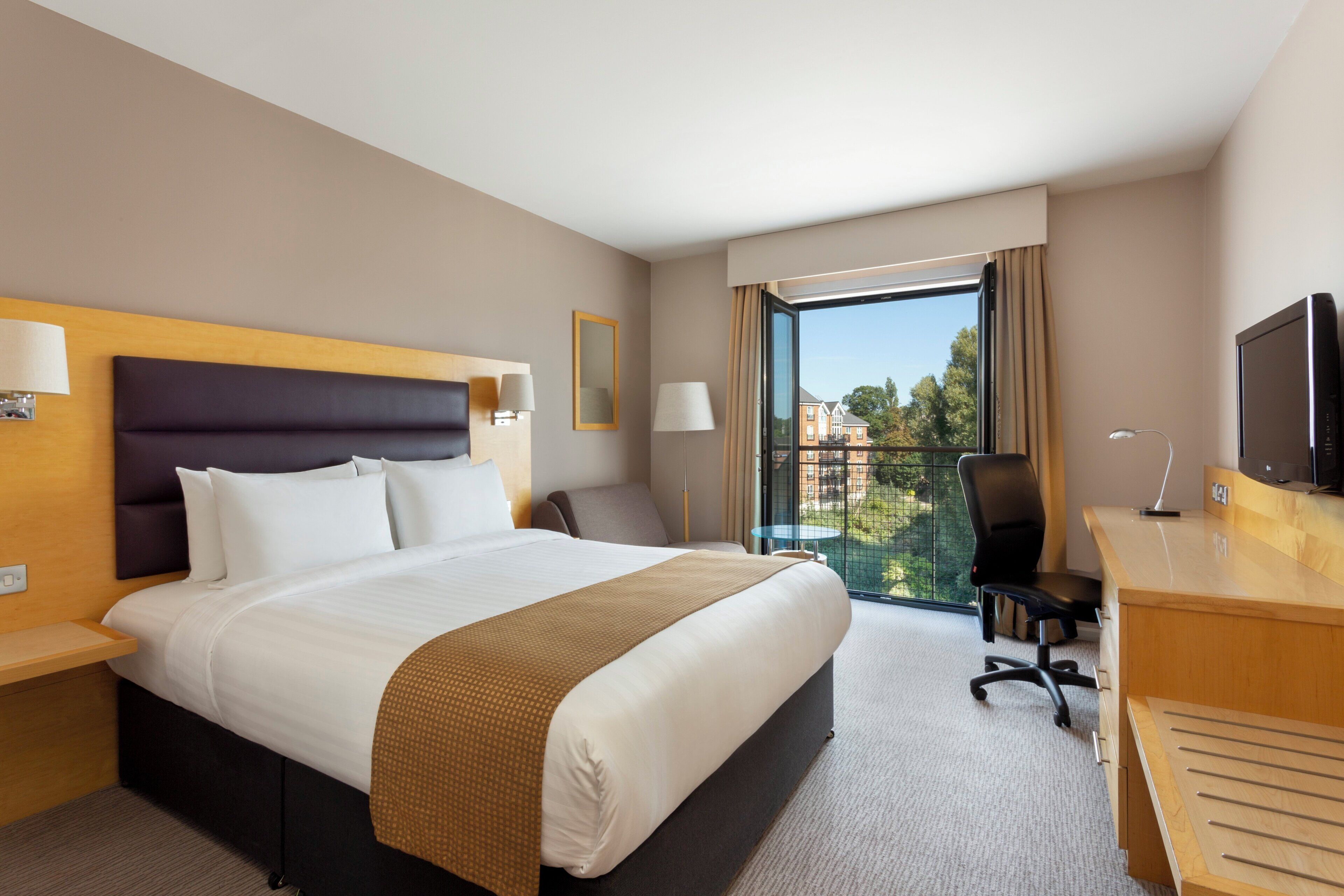 Une chambre d’hôtel avec un grand lit, un bureau, une chaise, une télévision et une fenêtre donnant sur des immeubles.