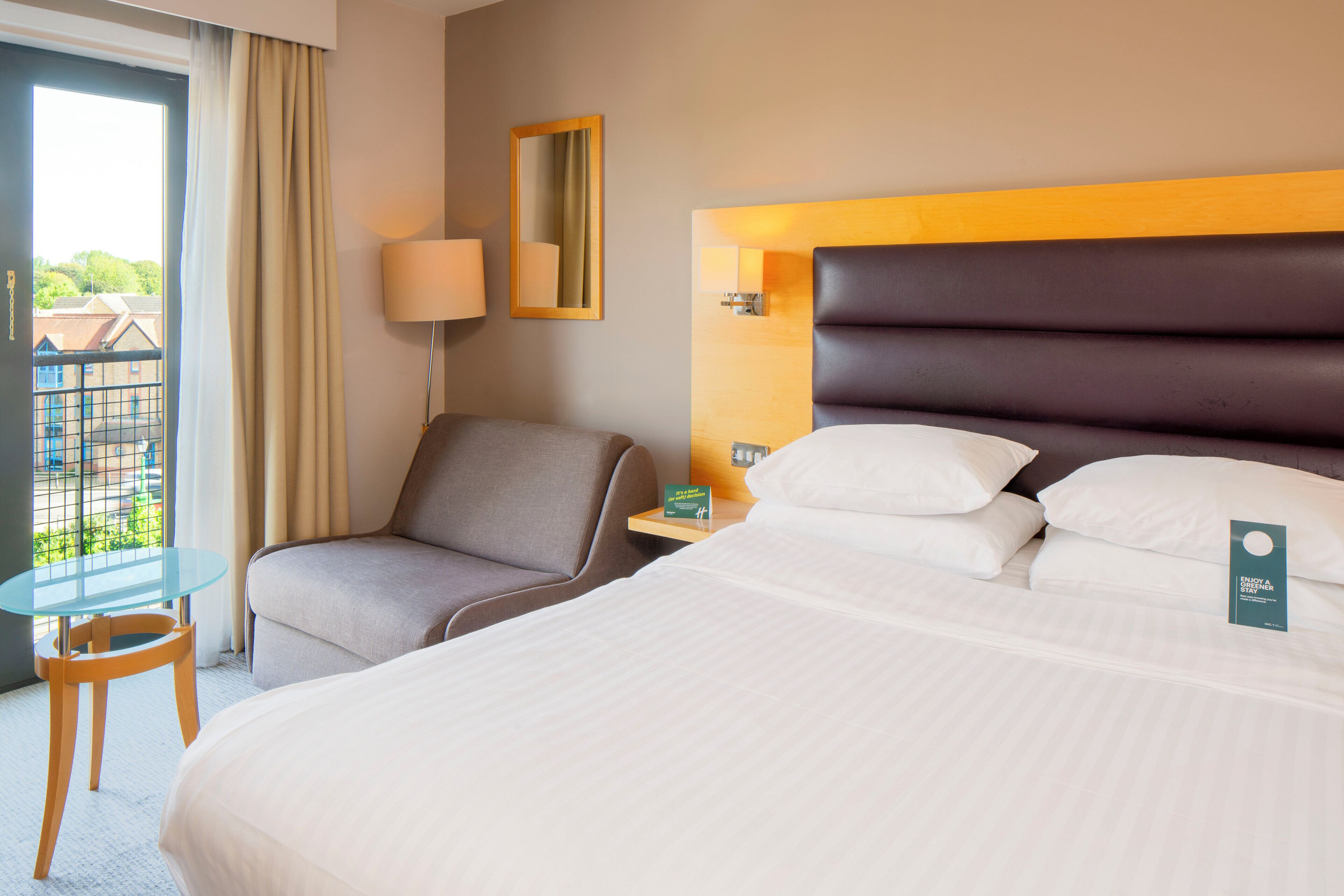 A hotel room with a large bed, a chair, a small table, a lamp, and a view of the outside.