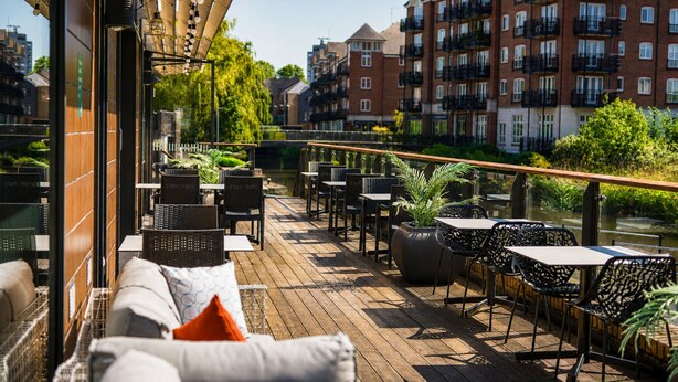 An outdoor deck with wicker furniture and potted plants, overlooking a river and buildings.