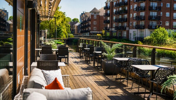 An outdoor deck with wicker furniture and potted plants, overlooking a river and buildings.