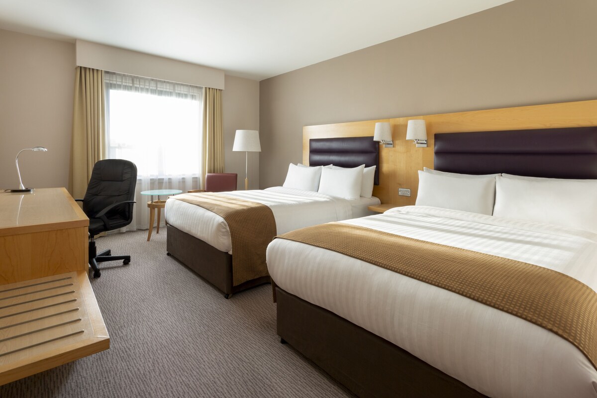 Ein Hotelzimmer mit zwei Betten, einem Schreibtisch, einem Stuhl, einer Lampe und einem Fenster mit Vorhängen.