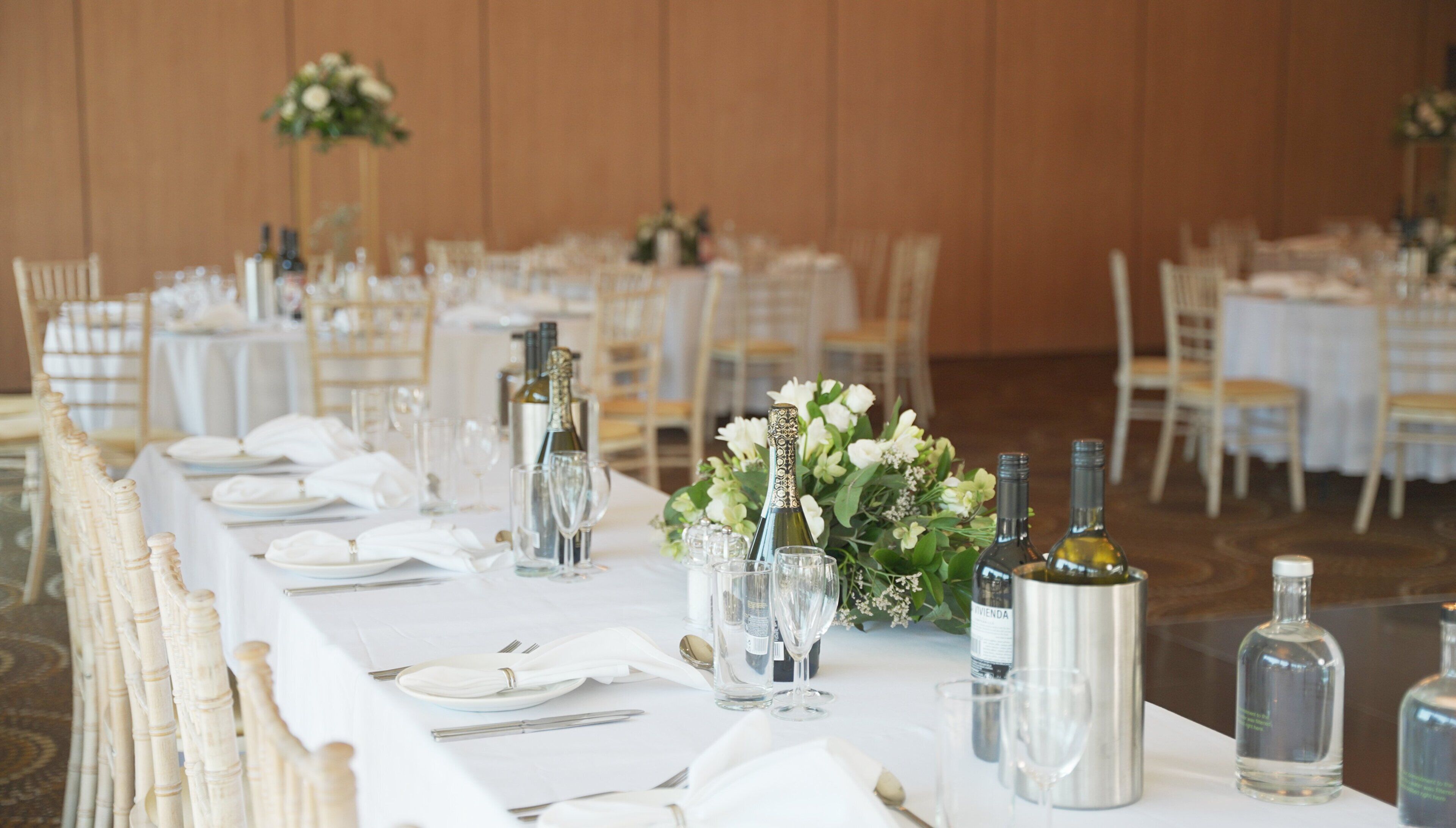 Un salón de banquetes con mesas redondas dispuestas para un evento formal, con manteles blancos, cristalería y centros de mesa florales.