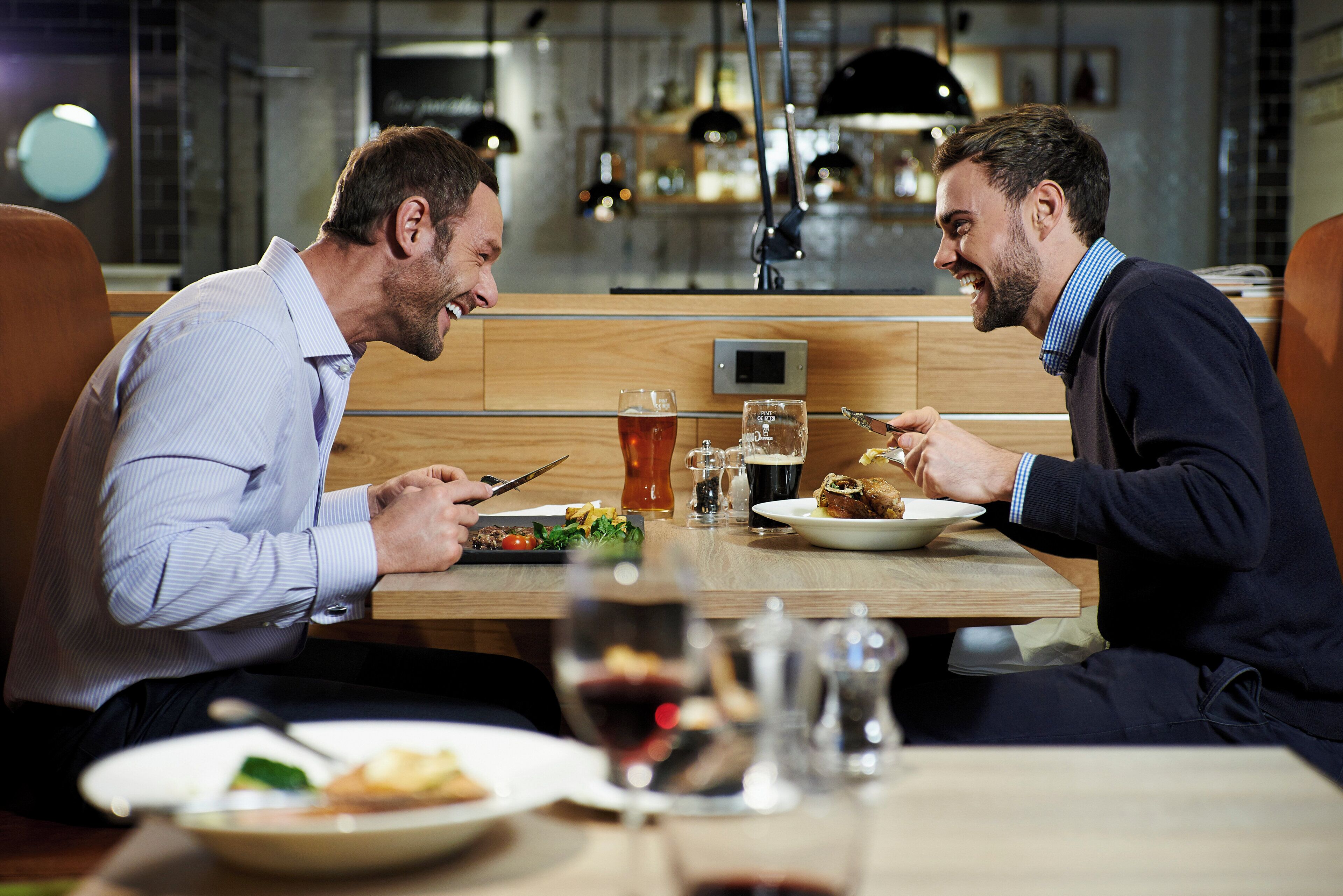Duas pessoas jantando em um restaurante, com uma mesa de madeira, pratos de comida e copos com bebidas.