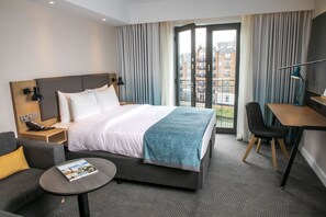 A modern hotel room with a large bed, a desk, a chair, and a view of buildings through the window.