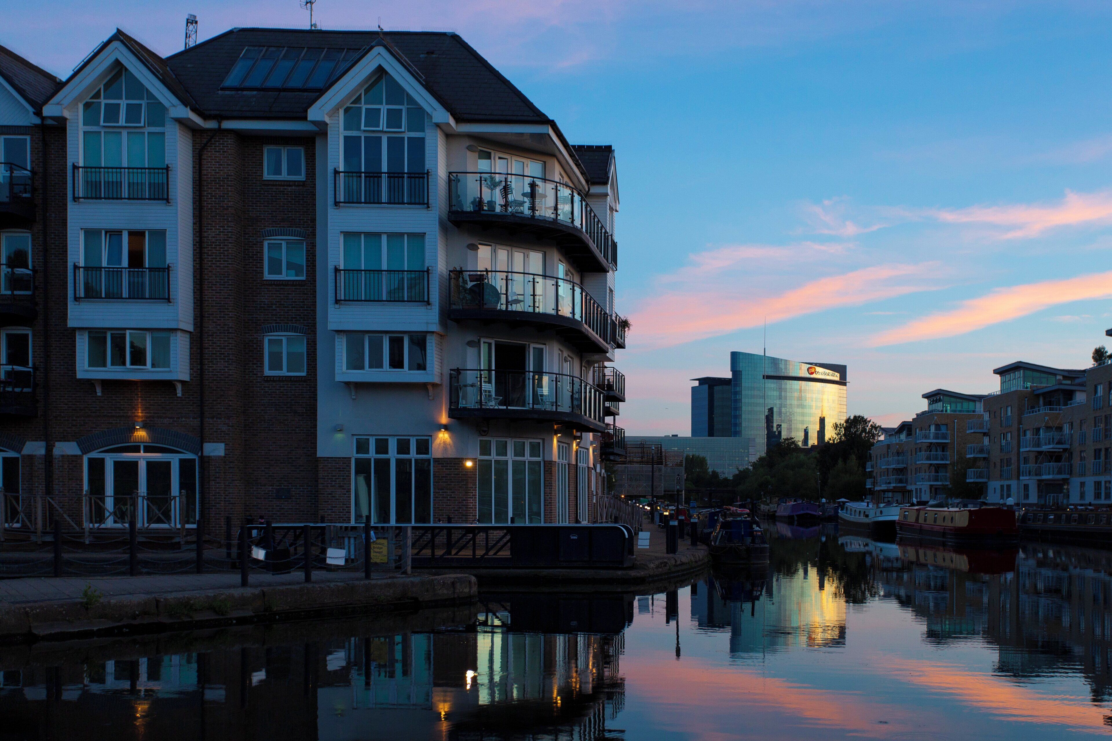 A multi-story residential building with balconies, a reflective water body in front, and a modern glass building in the background.