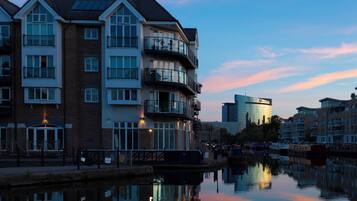 A multi-story residential building with balconies, a reflective water body in front, and a modern glass building in the background.
