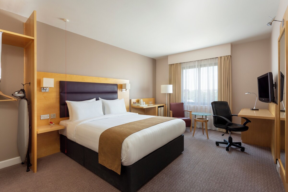 Ein Hotelzimmer mit Bett, Schreibtisch, Stuhl, Fernseher und einem Fenster mit Vorhängen.