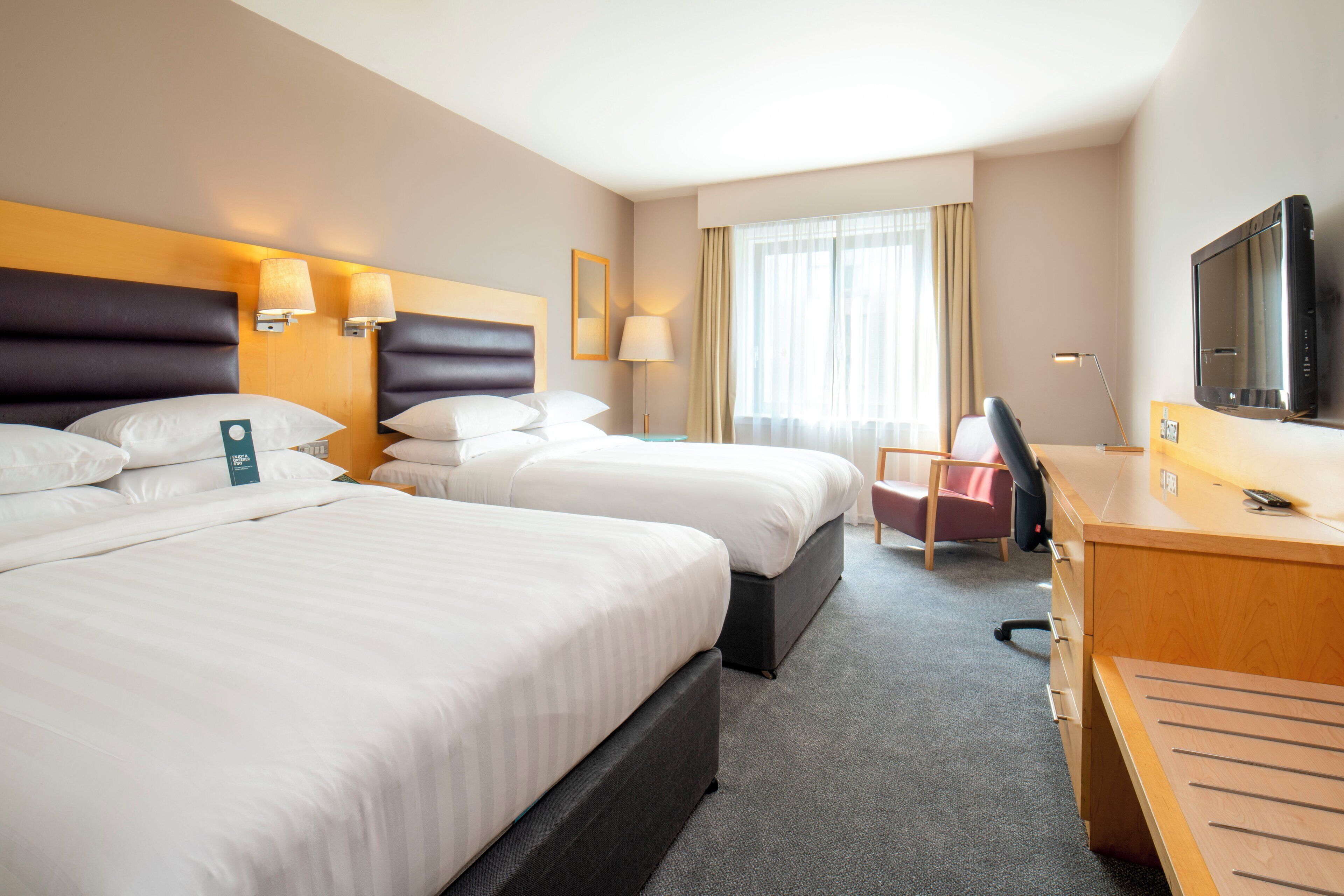A hotel room with two beds, a desk, a chair, a TV, and a window with curtains.