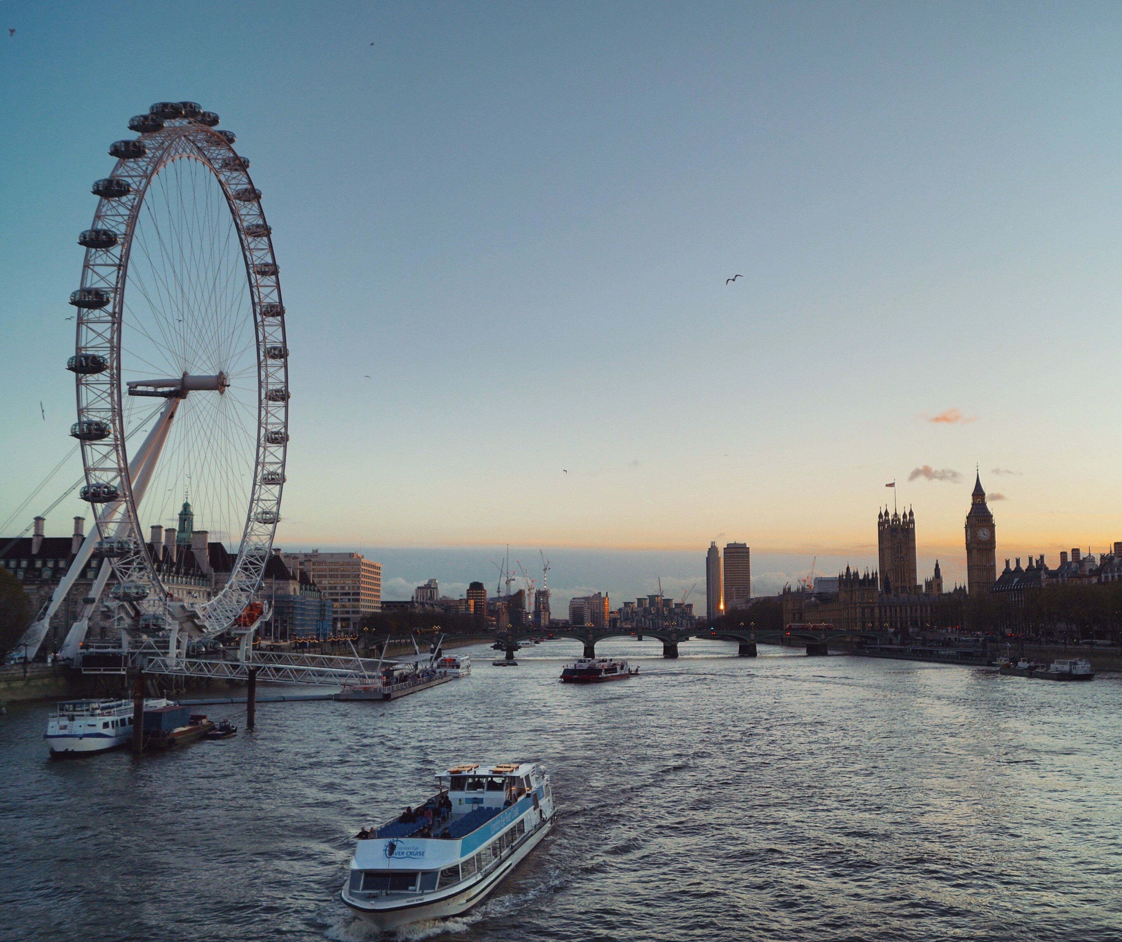 A river with boats, a large Ferris wheel, and the silhouette of a city skyline including a prominent tower.