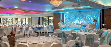 A banquet hall with round tables, chairs with blue sashes, chandeliers, and a stage with draped curtains.