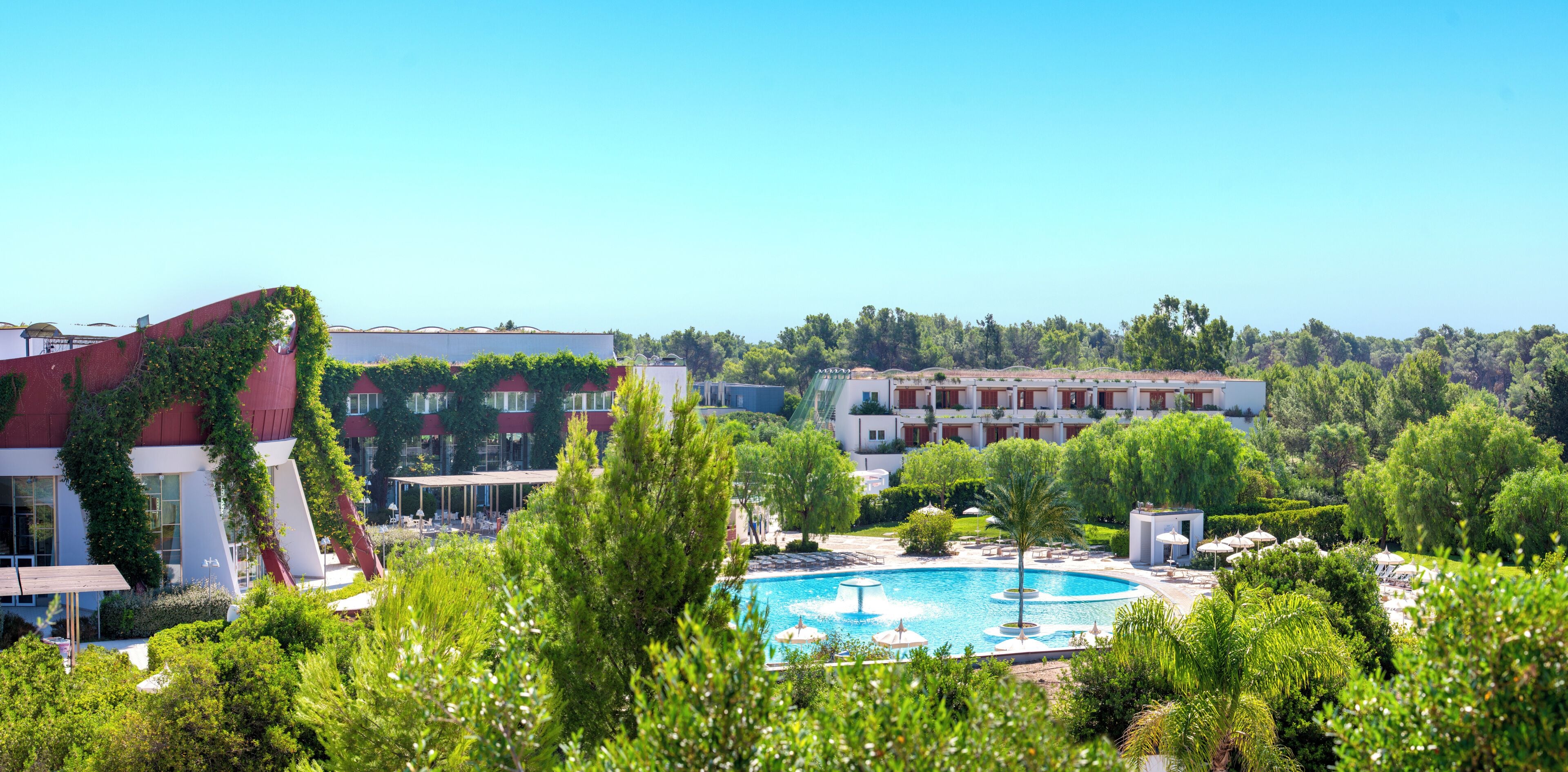 Un hotel con piscina rodeado de vegetación, un edificio rojo cubierto de hiedra y un edificio blanco con balcones.