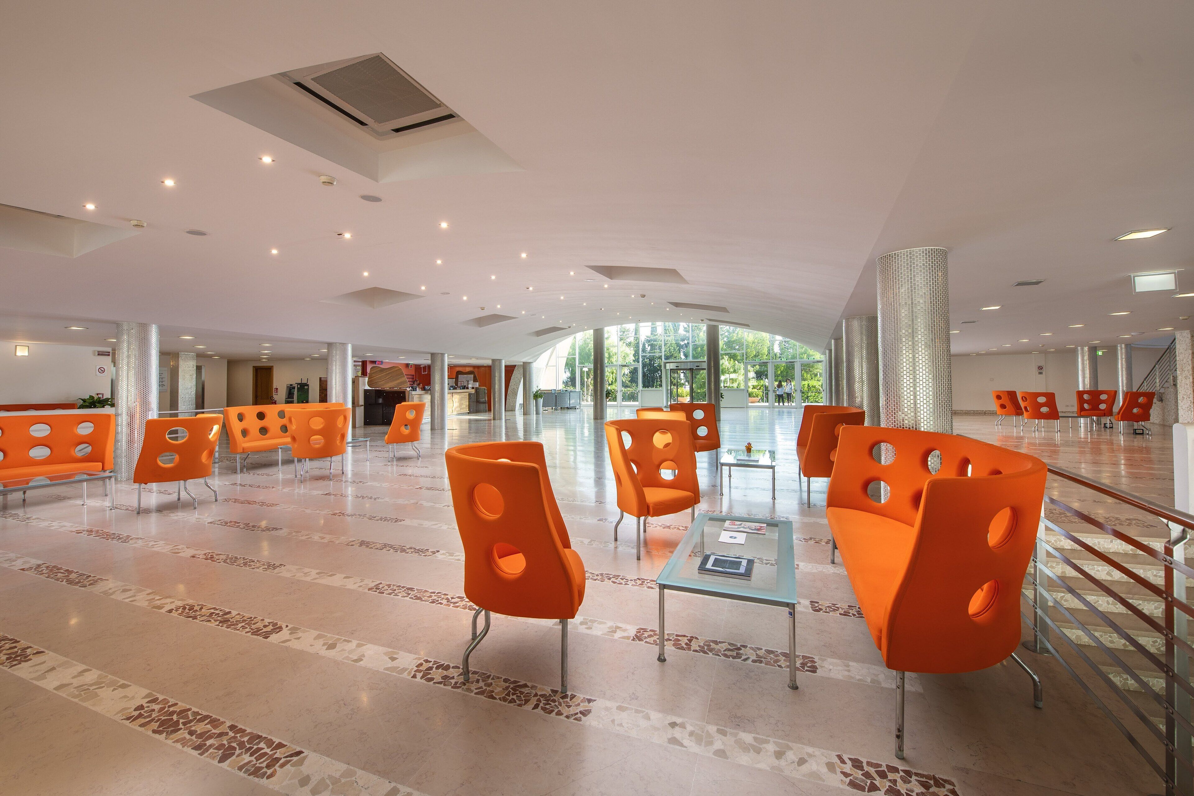 A spacious lobby with modern orange chairs, a glass table, and a view of greenery through large windows.