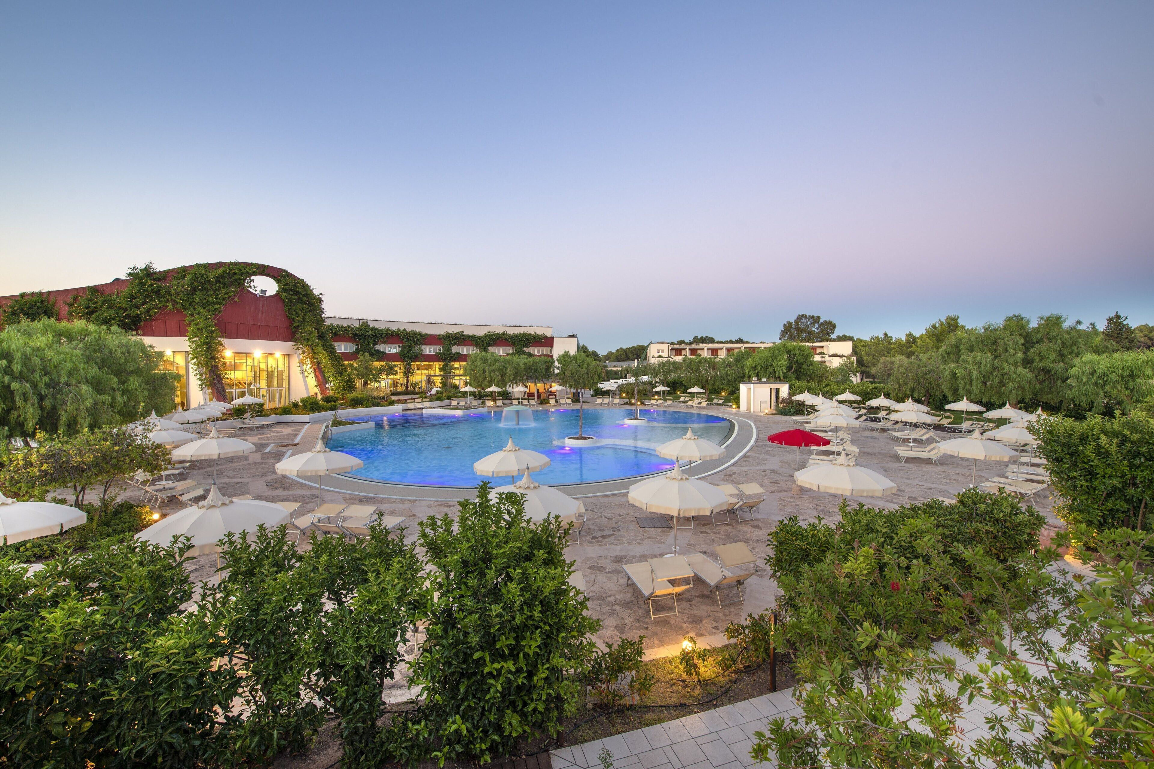 A large outdoor swimming pool surrounded by lounge chairs and umbrellas, with a building featuring a red roof and greenery in the background.