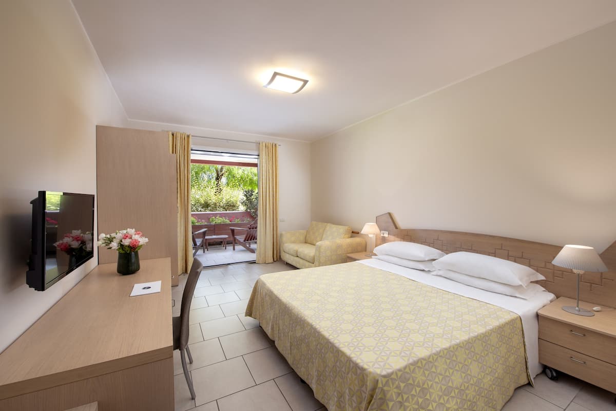 Ein Hotelzimmer mit Bett, Nachttisch, Lampe, Fernseher und einem kleinen Schreibtisch mit Blumenvase.