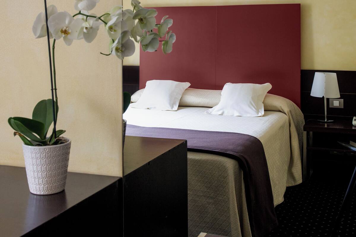 A hotel room with a bed, a desk, a laptop, a potted orchid, and a nightstand with a lamp.