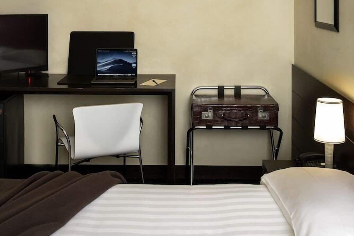 A hotel room with a bed, a desk with a laptop, a chair, a suitcase, a TV, and a lamp.