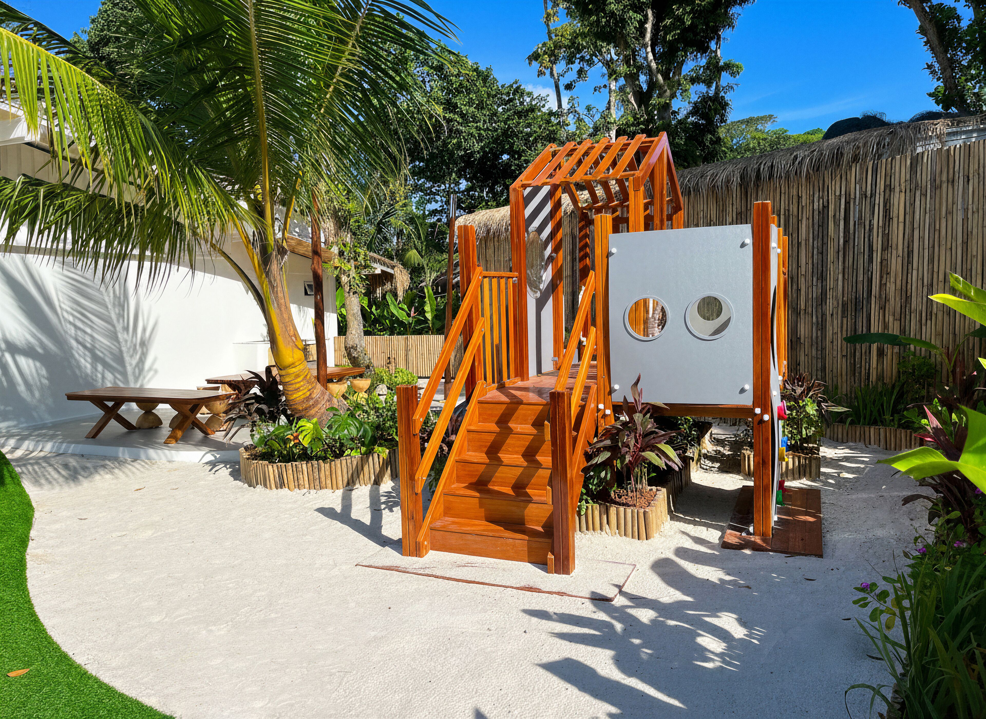 Een houten speelconstructie met een glijbaan en een klimmuur in een zandrijk buitengebied, met palmbomen en planten in potten.