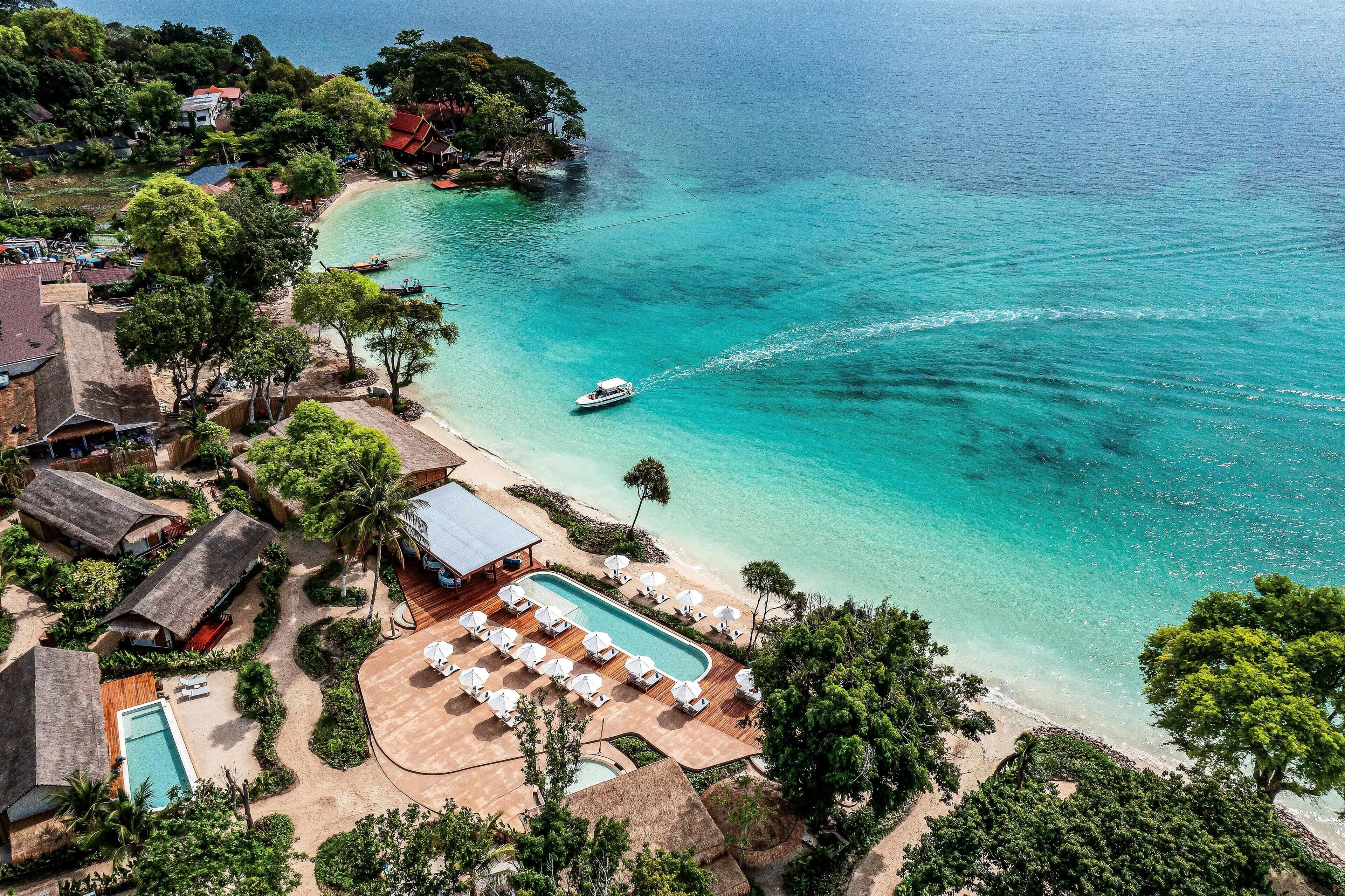 Vista aérea de un complejo turístico costero con piscina, playa y barcos.