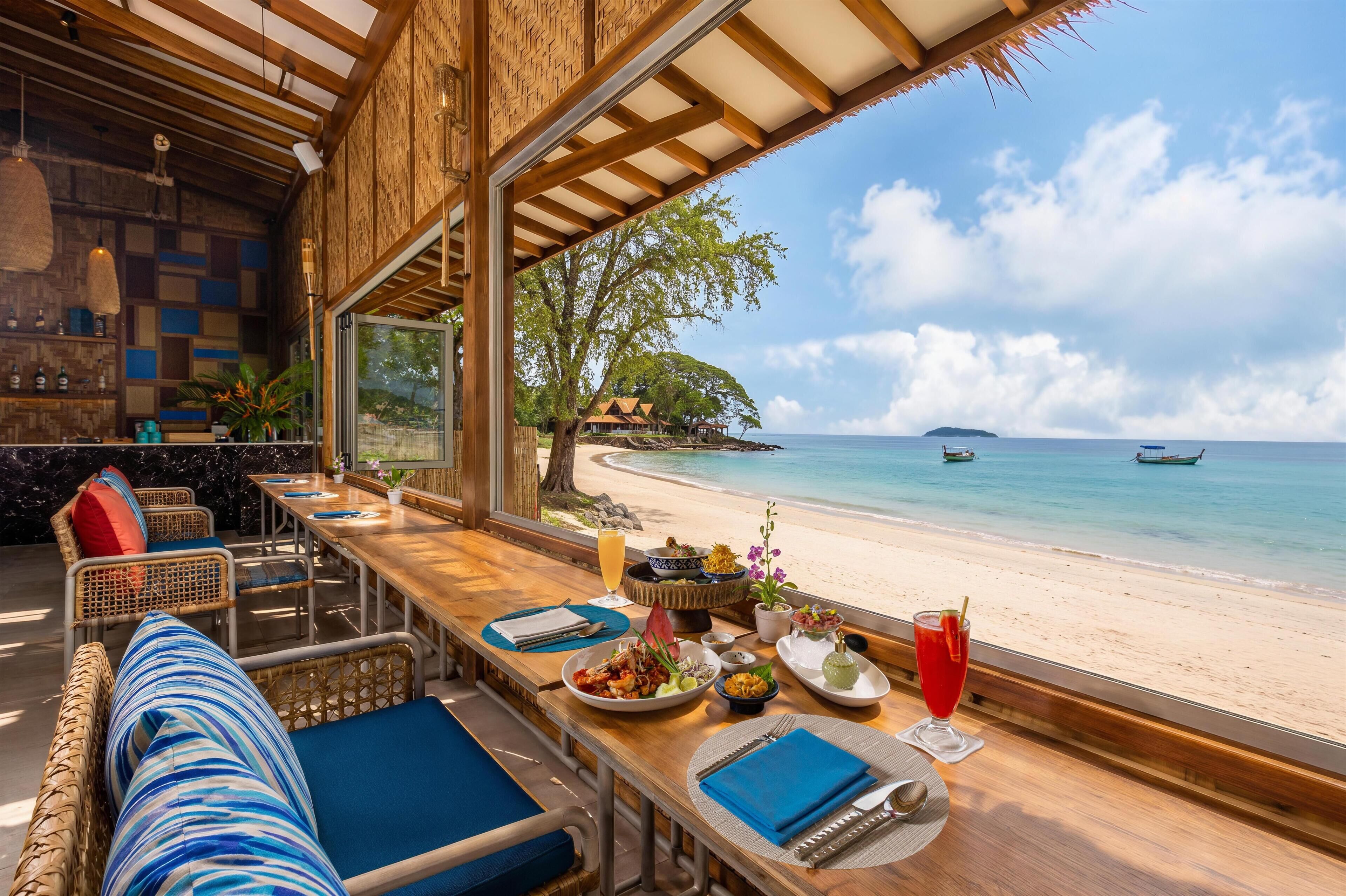 Un comedor con vistas a la playa y al océano, con una larga mesa de madera y sillas, un bufé con diferentes platos, y un ambiente tropical.