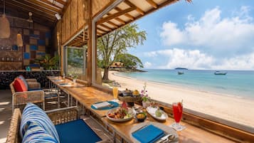 Un comedor con vistas a la playa y al océano, con una larga mesa de madera y sillas, un bufé con diferentes platos, y un ambiente tropical.